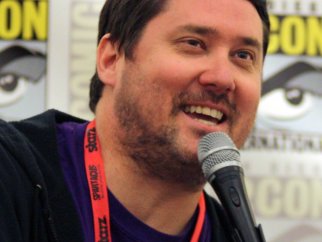 Comedian Doug Benson is holding a microphone and smiling.