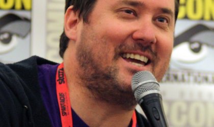 Comedian Doug Benson is holding a microphone and smiling.