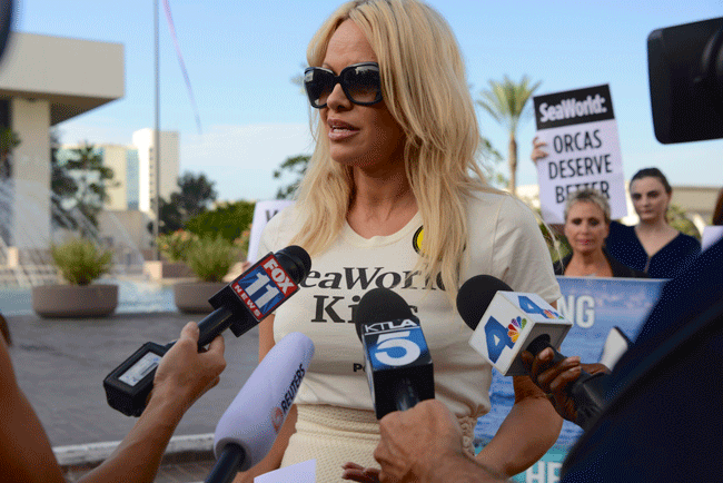 Pamela Anderson, a blond woman, holding a microphone in front of reporters.